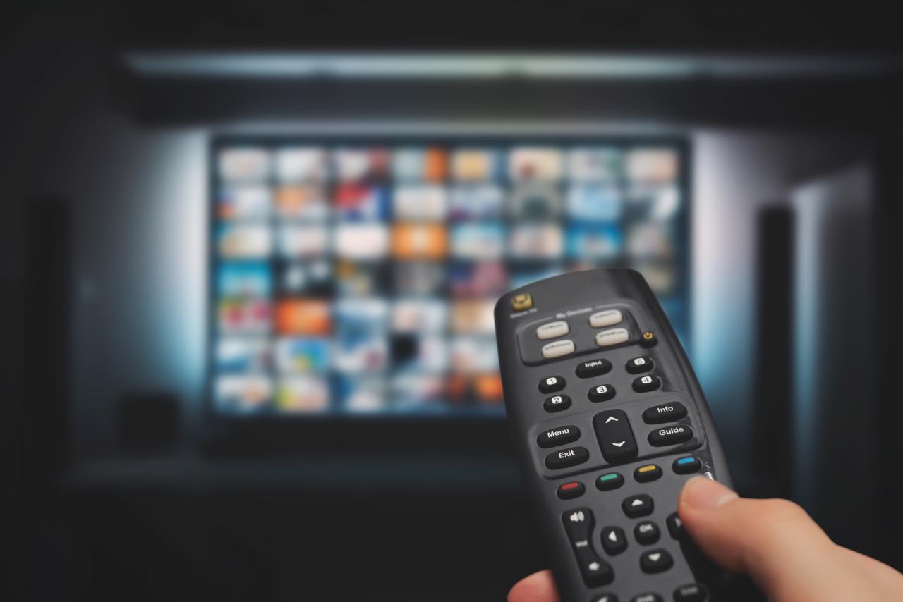 Hand Holding Remote Control In Front of Multichannel TV