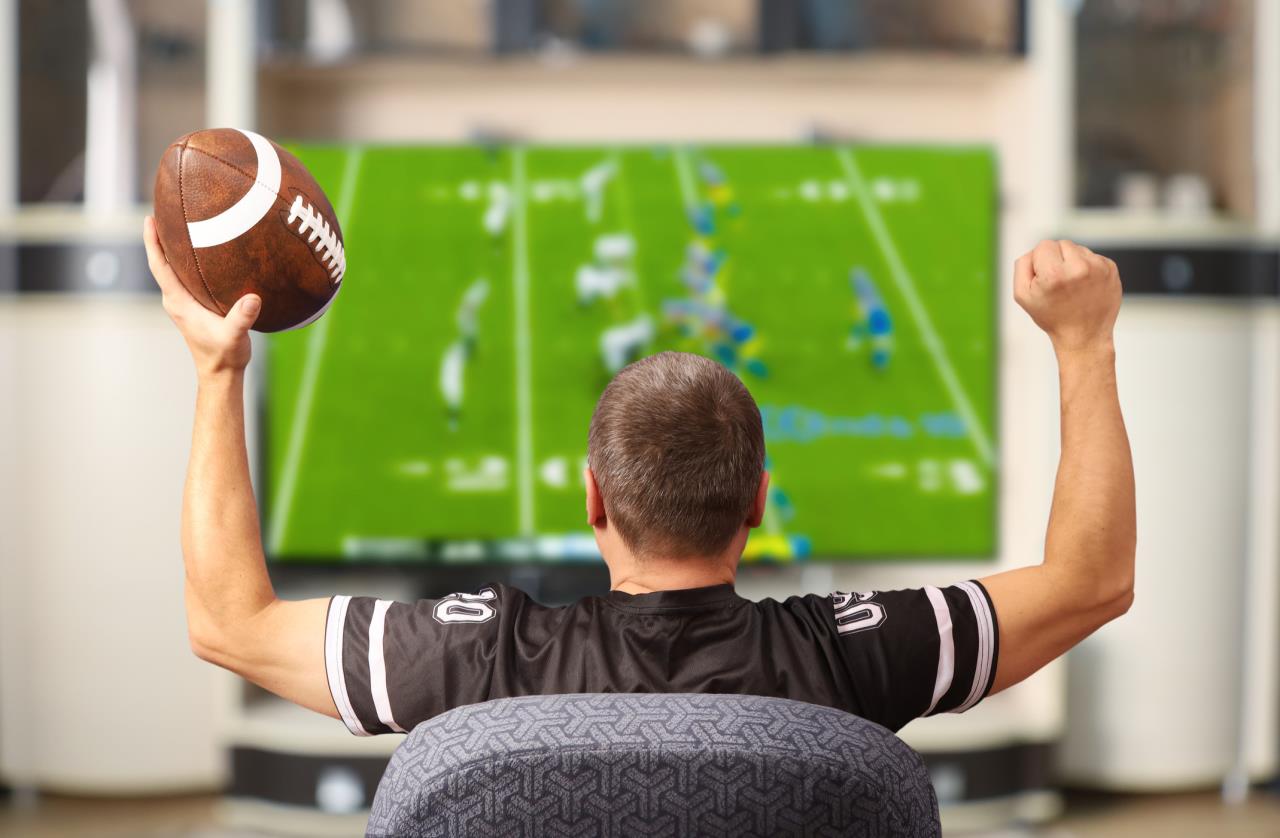 Man seen from behind watching football game on TV while raising football