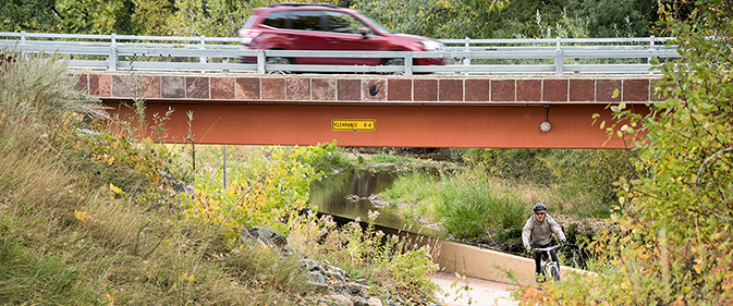 A bicyclist rides a riverside path under a bridge while a car passes overhead.