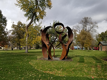 A tall contemporary art sculpture of thin rust brown ribbon like circles stacked one on top of each other on a green lawn with background trees.