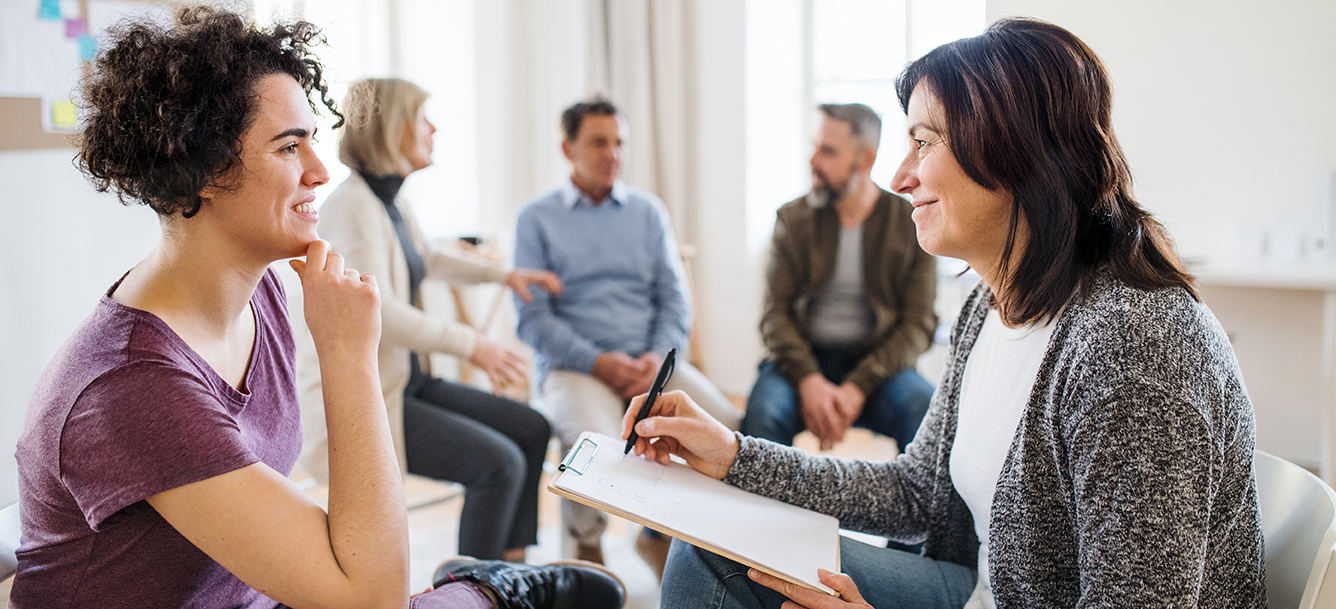 A senior counsellor with clipboard talking to a young woman during group therapy.