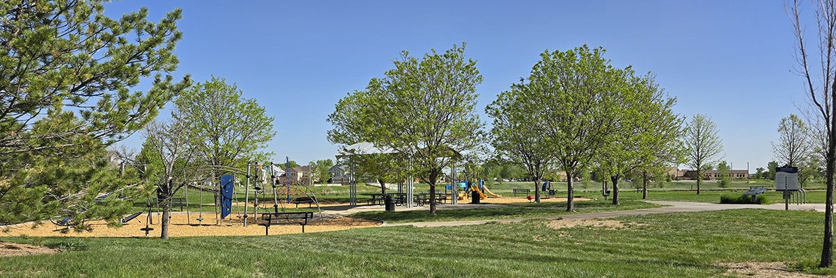 Green grass, trees and play structures are Blue Skies Neighborhood Park are seen against a blue sky.