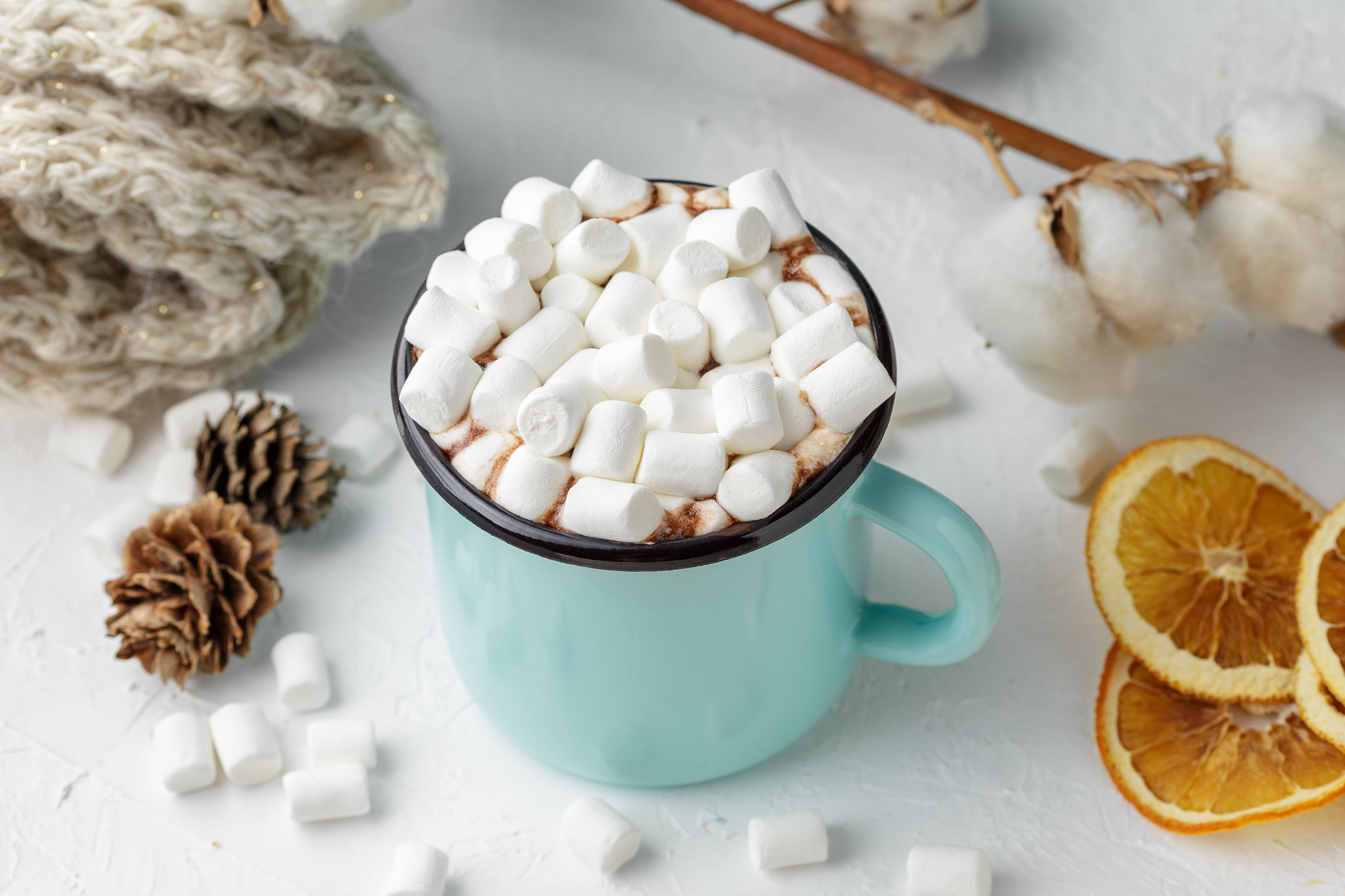 Blue metal mug of cocoa ot hot chocolate with marshmallows on the table with winter decor, branch of cotton flower, knitted blanket, dry citrus slices and wooden Christmas ornaments