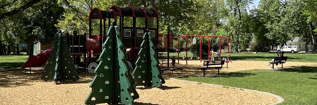 Collyer Neighborhood Park is pictured with some green pine tree-shaped playground structures in the foreground and a larger train-shaped play structure in the back ground.
