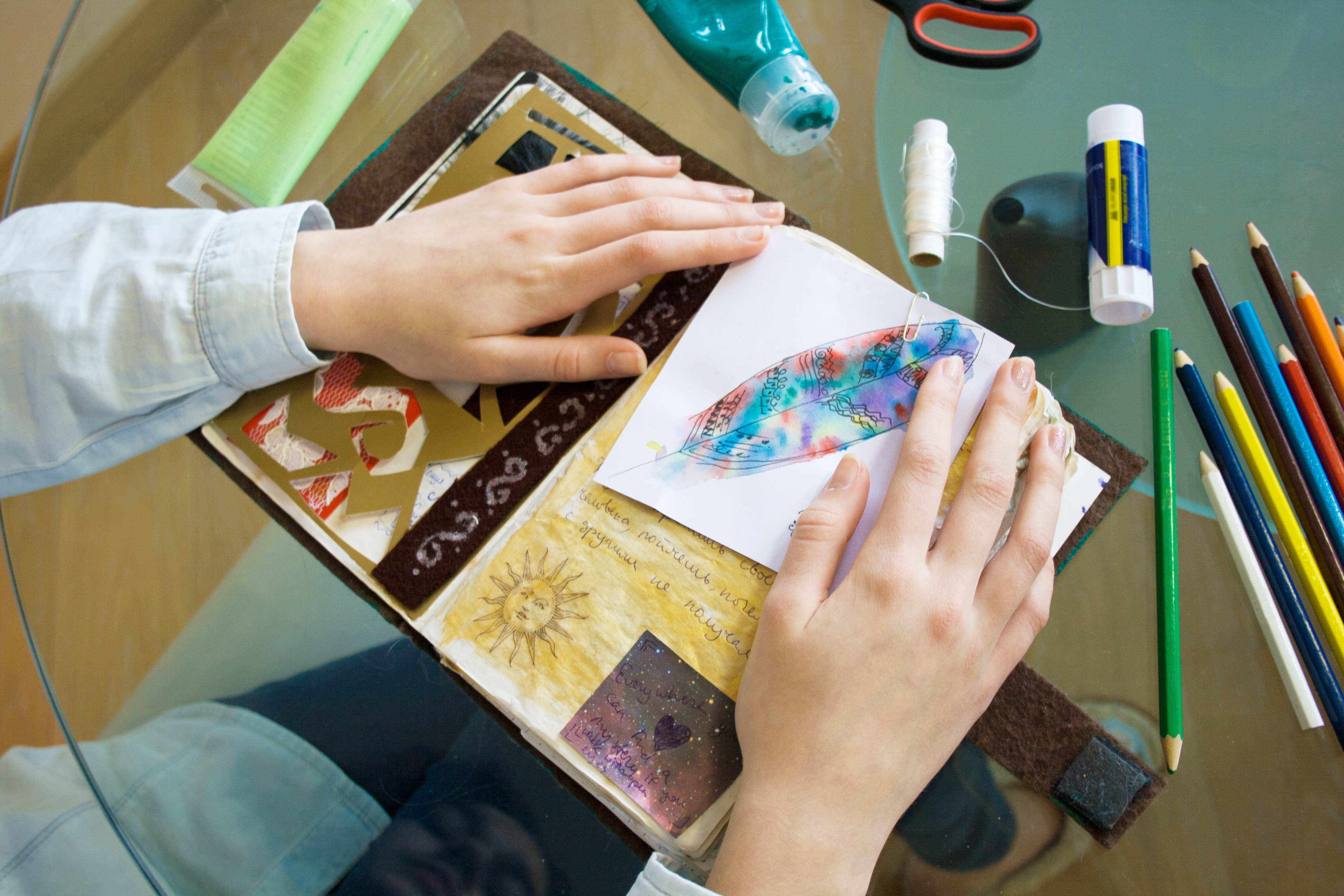 Table with elements for scrapbooking handcraft. Tools for decoration, handmade album, scissors, paint, glue, colored pencils, scotch, thread and hands of teenager girl master. Top view.