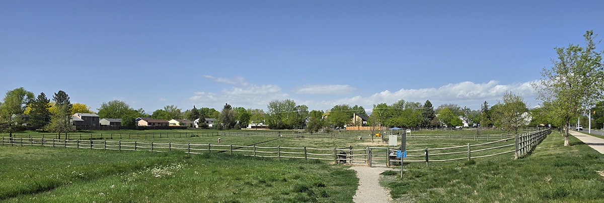 The fences and green grass of dog park number one is seen with a partly cloudy sky in the background.