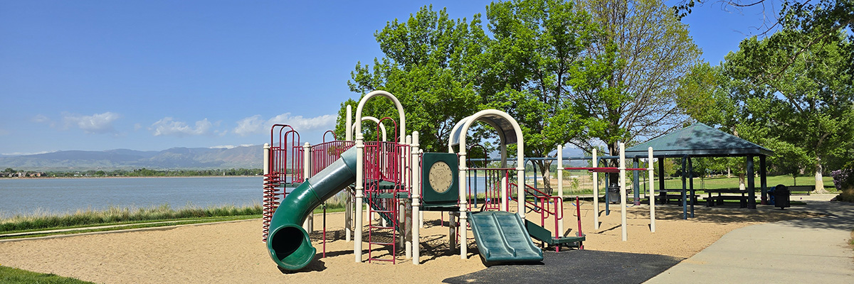 A colorful red and green play structure sits on white sand with McIntosh Lake and mountains in the background.