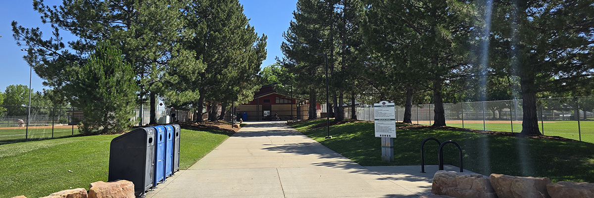 A tree-lined sidewalk cuts between two fenced sports fields at Longmont's Garden Acres Park.