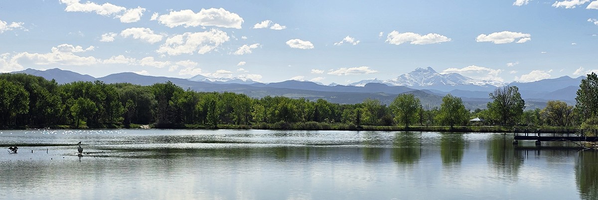 A view of the Golden Ponds Nature Area shows a beautiful pond surrounded by green trees with mountains and blue skies in the background.