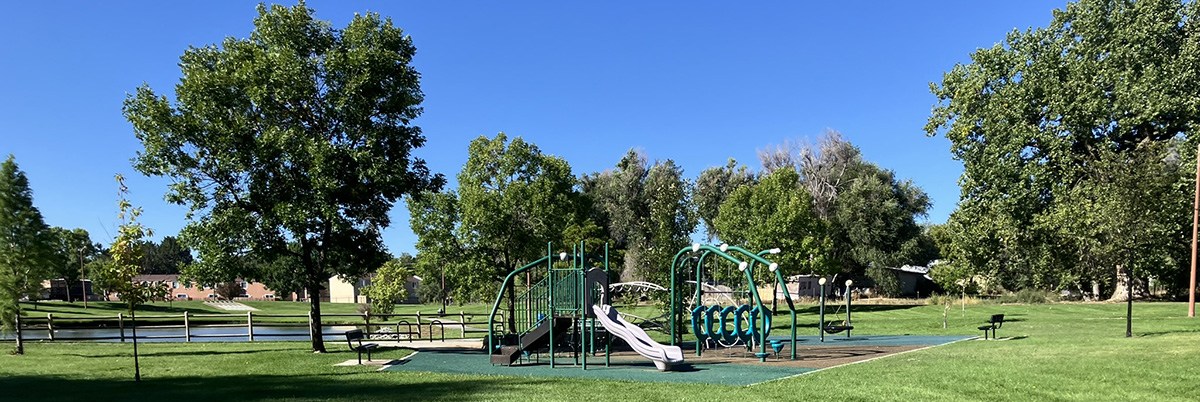 Longmont's Kensington Neighborhood Park is pictured with a colorful play structure surrounded by grass and leafy green trees.