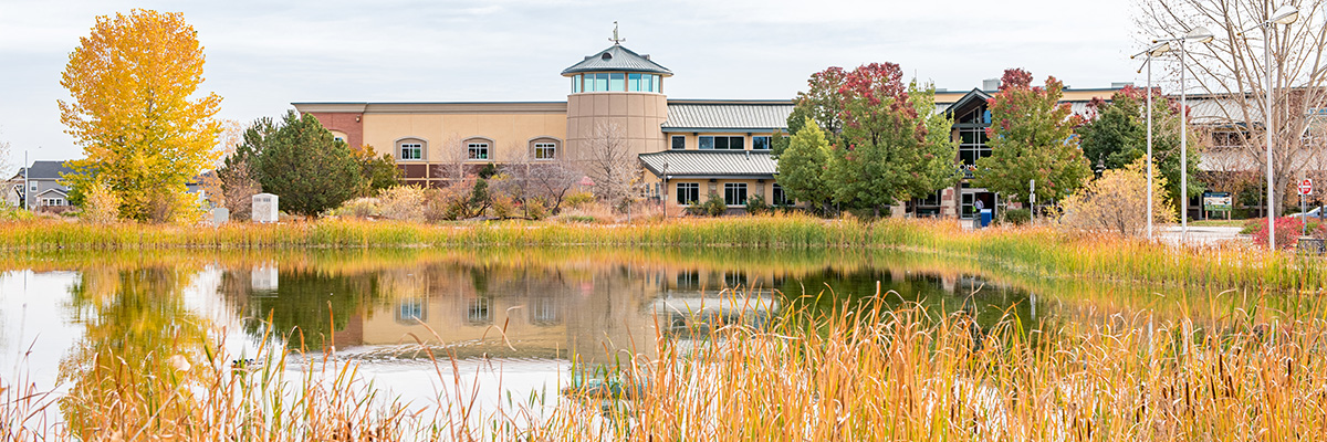 The Longmont Recreation Center is seen in autumn, surrounded by colorful leaves with a cattail-ringed pond in the foreground.