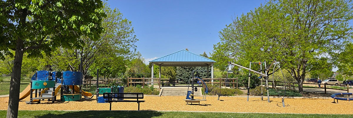 A new playground is seen in front of a park shelter, both surrounded by green grass and leafy trees.