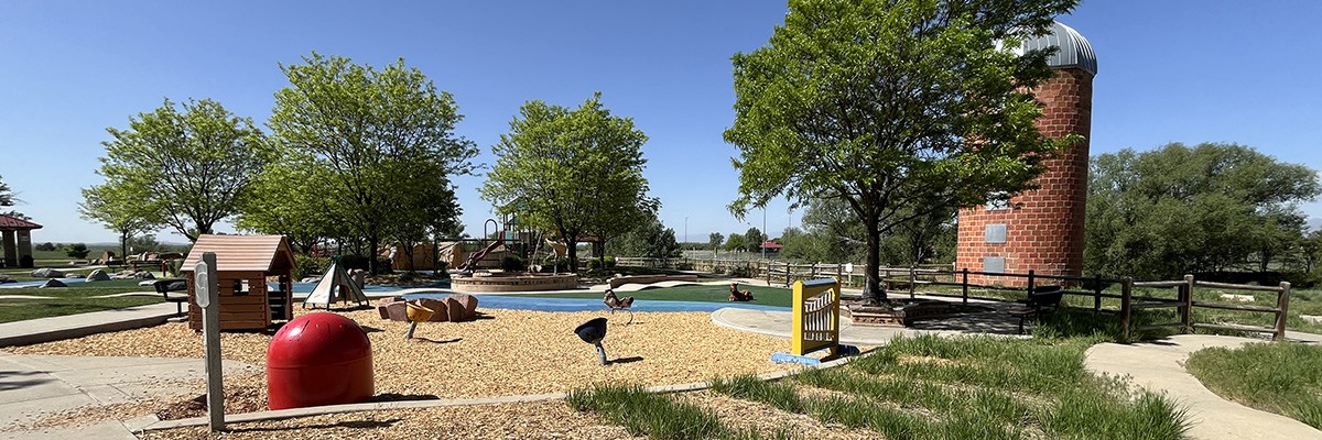 A playground and a brick silo are surrounded by green grass and leafy trees.