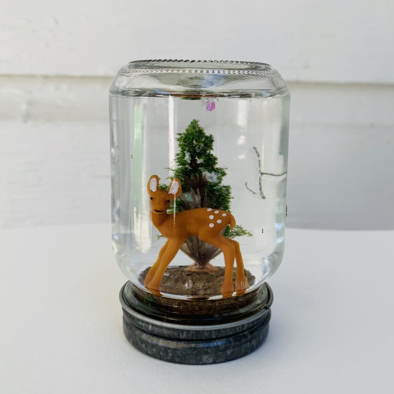 A mason jar snowglobe with a deer and a pine tree inside