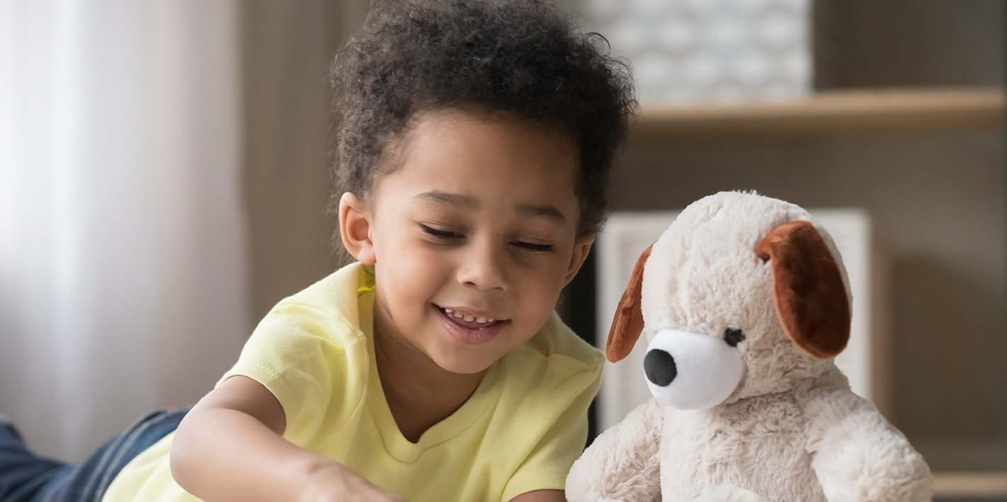 Happy mixed race little boy enjoying playing alone reading book to fluffy toy lying on warm floor, cute smiling african american kid having fun at home, creative child activity, underfloor heating