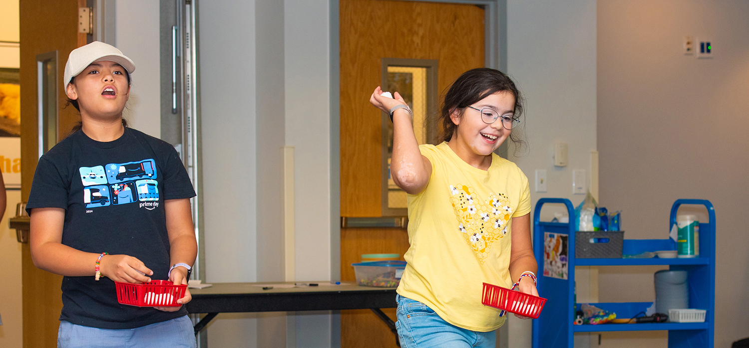 Two girls throw cotton balls into buckets during the Library's Tween Club