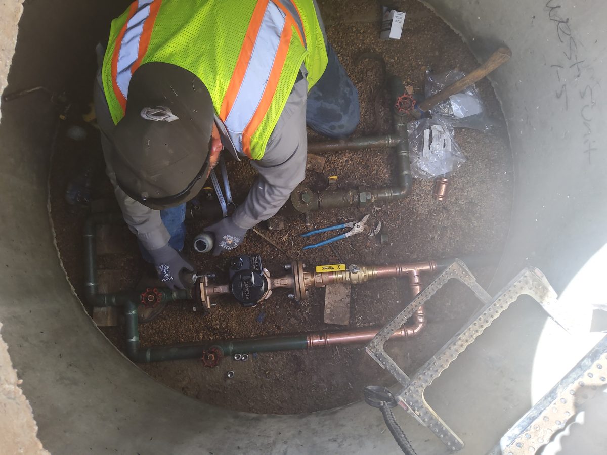 A Utilities and Public Works employee performs maintenance on a water meter, with tools and pipes close at hand.