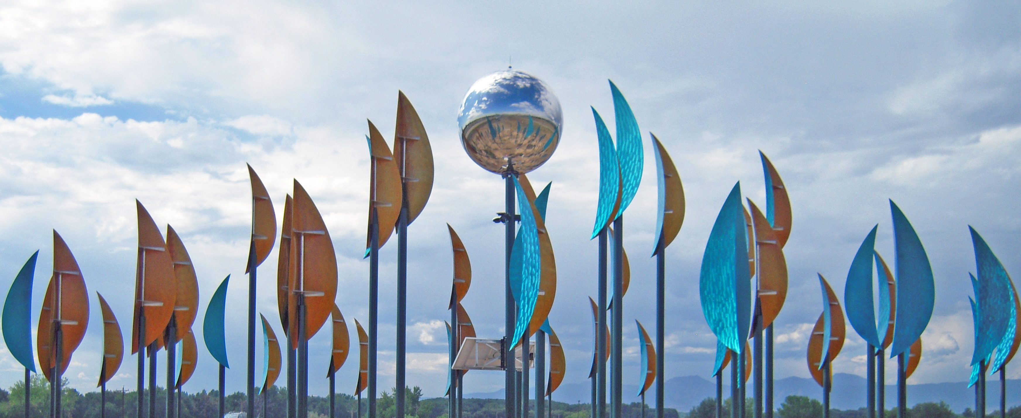 an outdoor public sculpture with abstract leaves and a shiny orb with a cloudy sky.