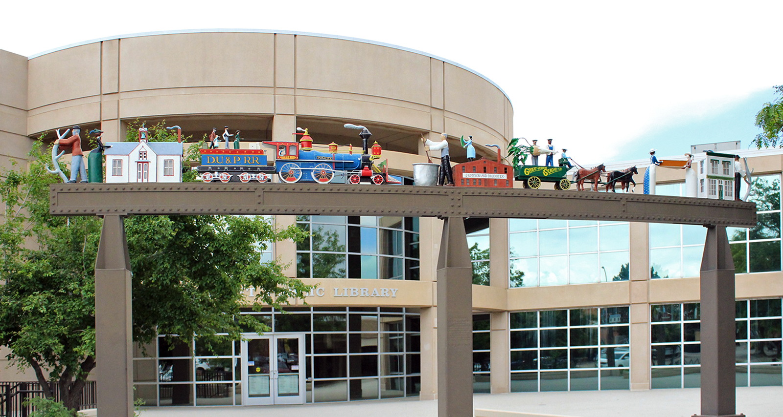 a sculpture with various people, buildings, animals and modes of transportation from the late 1800's on a steel entryway in front of the Longmont Library