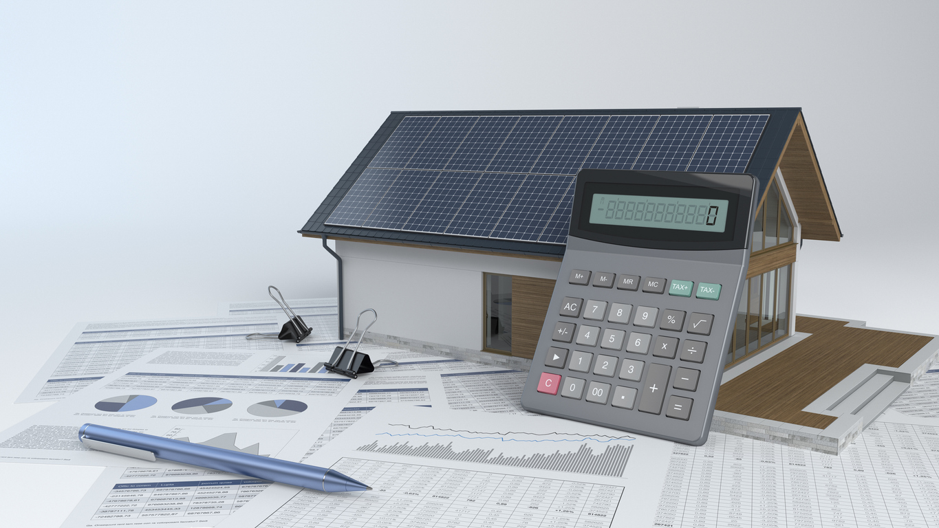 Calculator leaning against model of house with rooftop solar panels, with pen and financial documents nearby