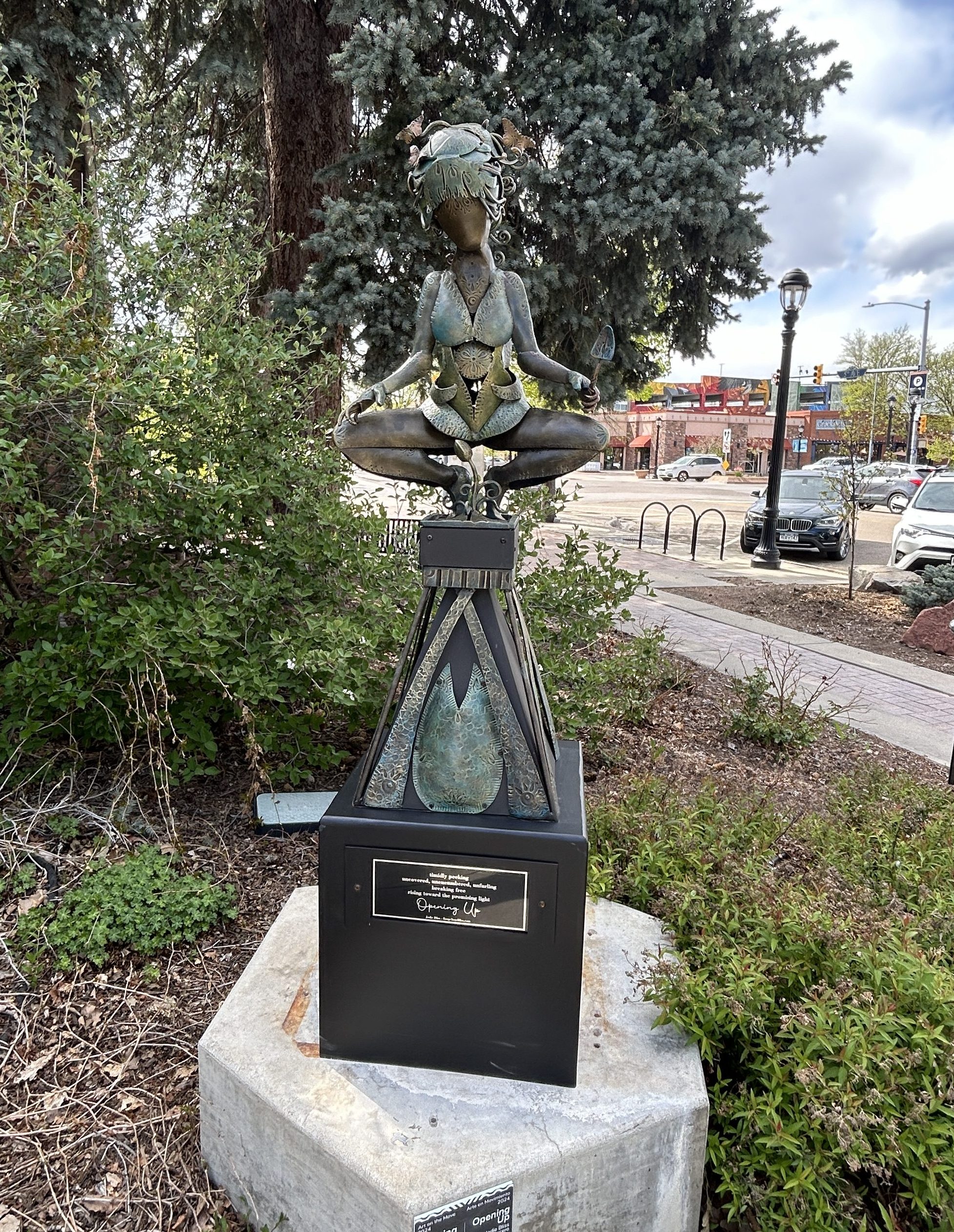 a sculpture of a woman above a small plant in hammered steel pictured in a park