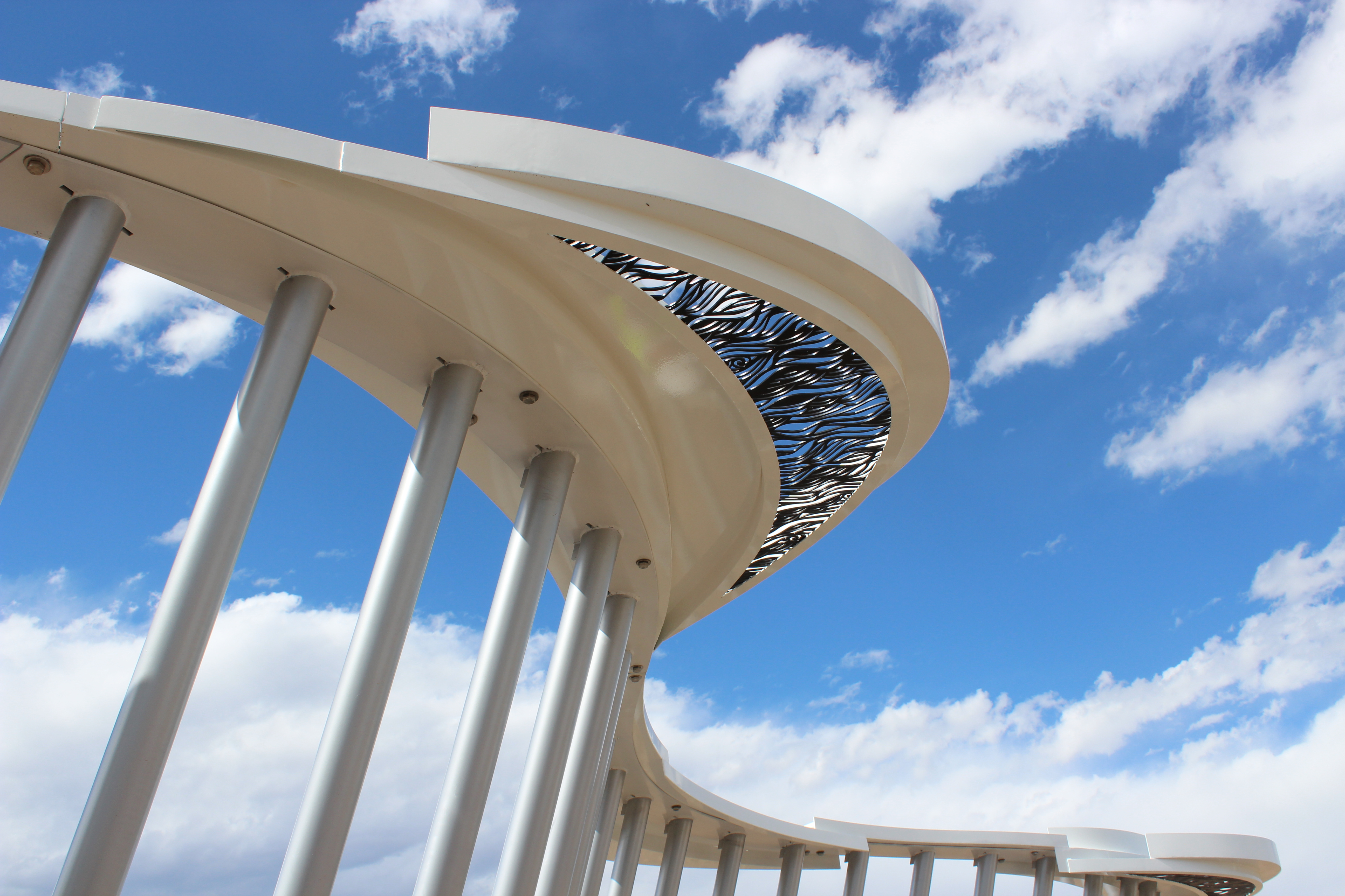 an abstract sculpture with metal poles and a wavy awning in front of a sky with clouds