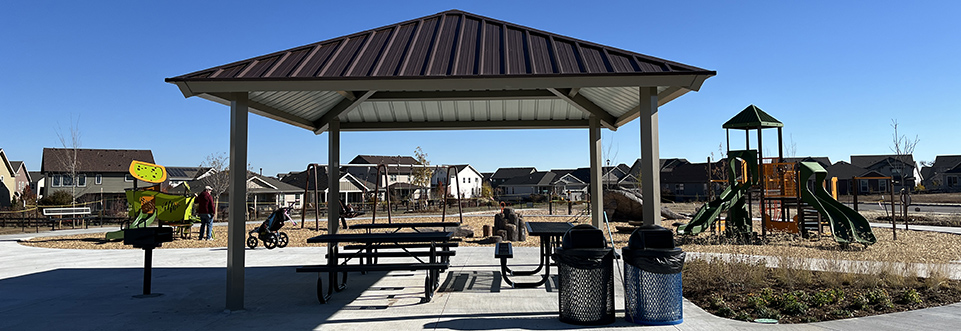 Clover Meadows Park is seen on a sunny, cloudless day. A picnic shelter is seen with a play structure in the distance.