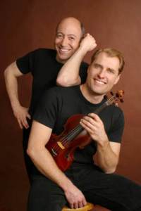 Two men in black shirts one holding a violin against a brown backdrop 