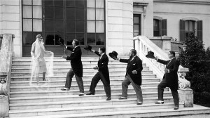 Black and white film still of several men in black staggered up the stairs outside a building