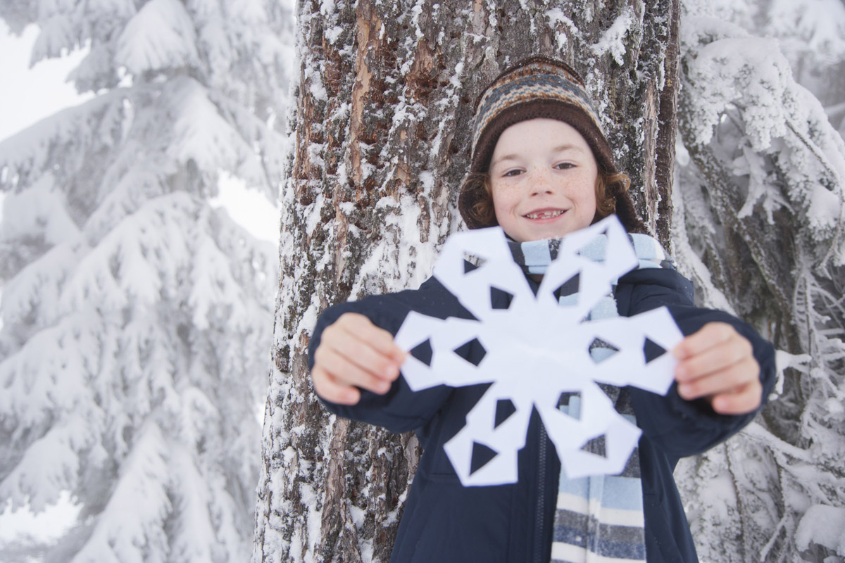 child in coat and hat next to tree in winter holding paper snowflake