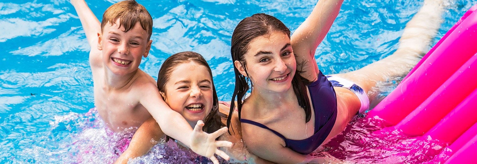 group of three kids in swimming pool with arms raised and a bright pink pool float