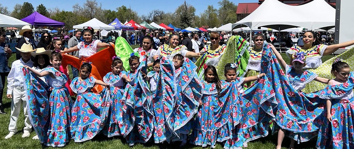 Many young girls are wearing traditional dance costumes while preparing to dance at the Longmont Cinco de Mayo festival. They are accompanied by women in traditional dance costumes and one or two young boys with cowboy hats.