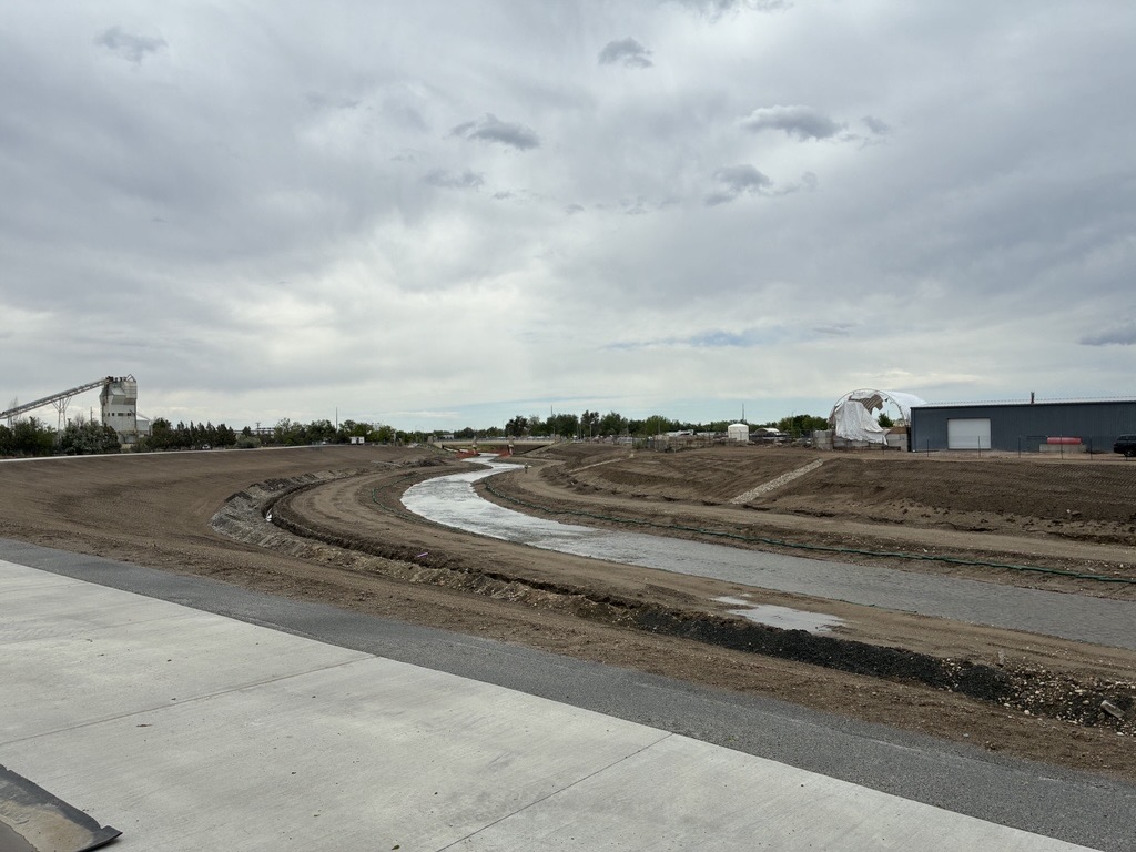 A calm and narrow creek winding through a dirt-covered landscape with a cement path in the lower left corner.