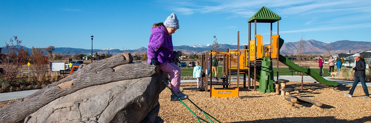 Clover Meadows Neighborhood Park is pictured with a young girl on a rock sculpture in the foreground, a green and yellow play structure in the middle ground, and the mountains and blue sky in the background.