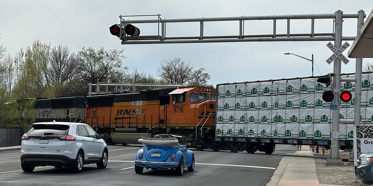 Orange Train crossing 9th Avenue in Longmont with a white SUV and Blue car waiting.