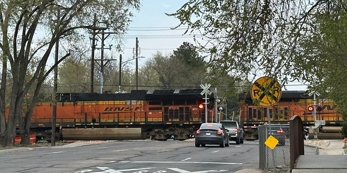 Orange Train crossing 17th Avenue in Longmont with three gray vehicles waiting and yellow railroad crossing signs.