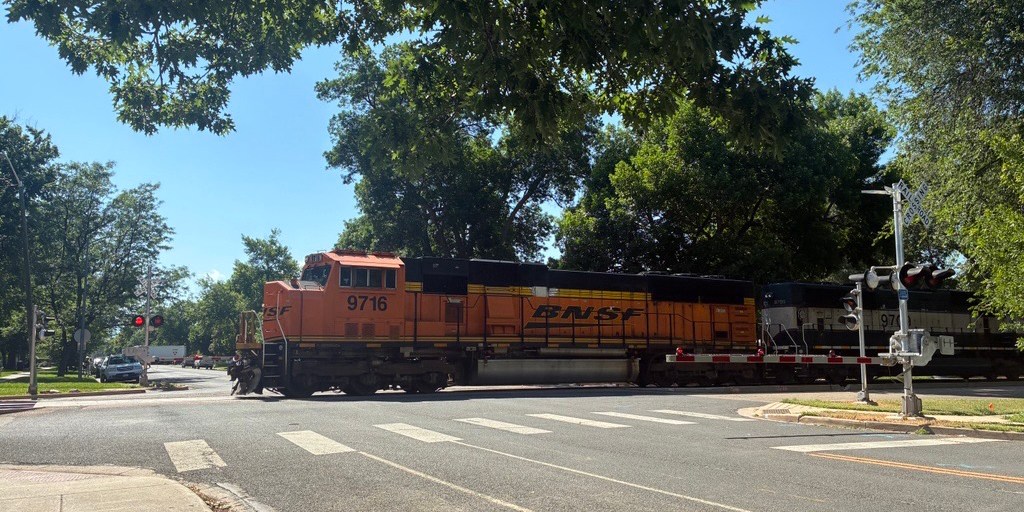 Orange Train crossing Longs Peak Avenue in Longmont with green trees in the background under a blue sky.