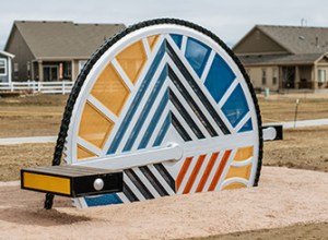 An oversized, colorful bicycle spoke wedged into the ground with two benches that look like bicycle pedals sits in a bed of red gravel.