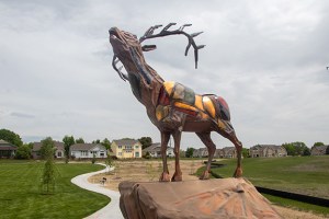 A bugling elk sculpture comprised of colorful sheet metal stand on top of a rock with green grass in the background