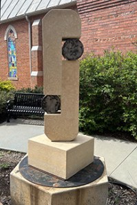 Cream colored limestone serpentine sculpture with two metal drums on either side sitting on a concrete pedestal. In the background is a brick building and green bushes.