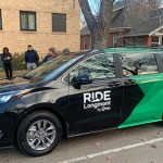 A black and green RIDE Longmont van is pulled up to a curb where riders await.