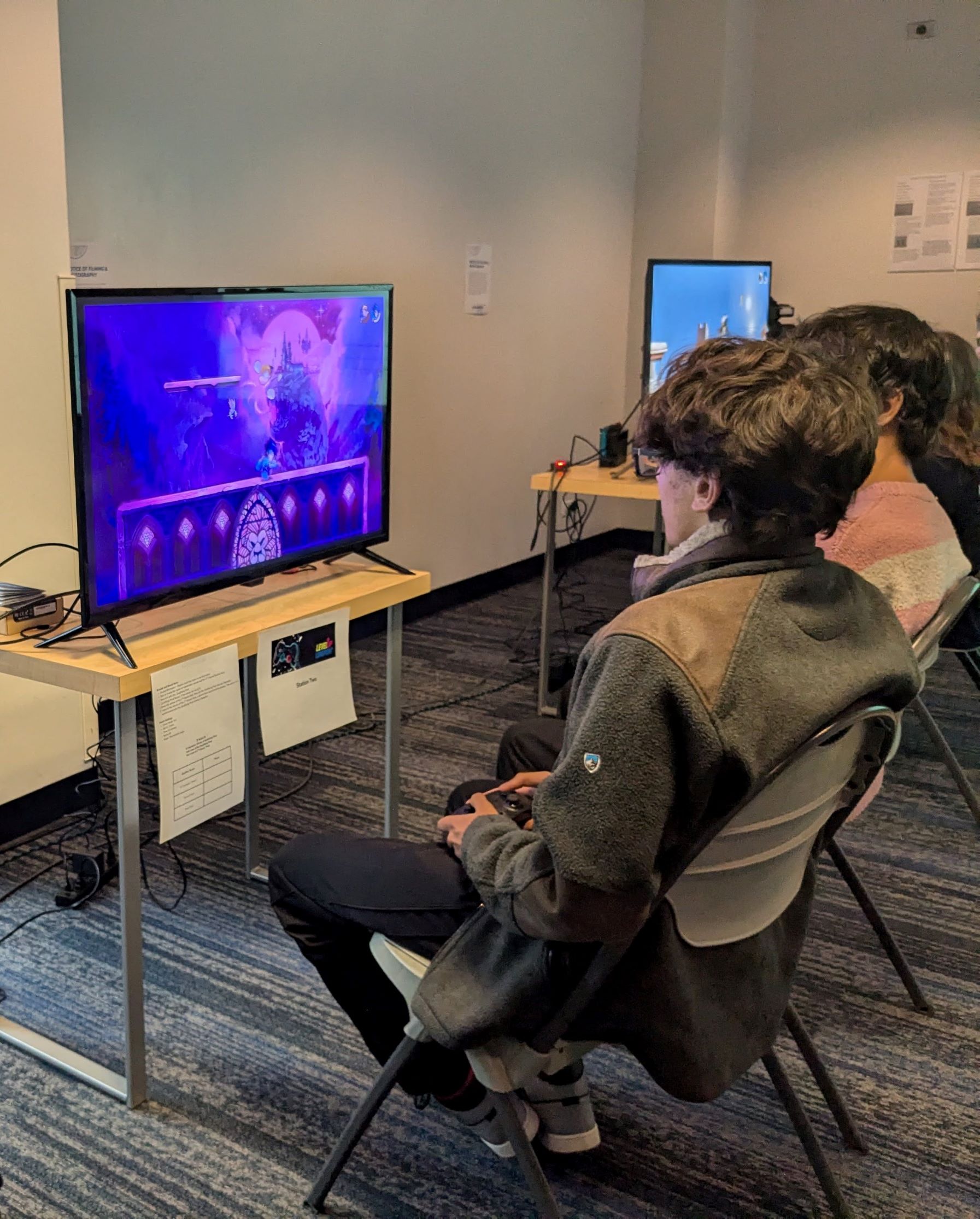 Two video gamers seated in folding chairs watch the action on a television screen or monitor in front of them.