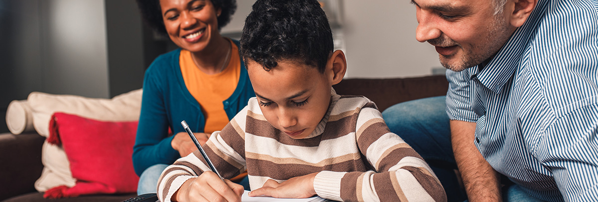 Two parents watch as a young child writes on a page.