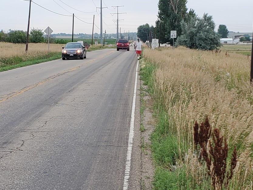 A black car driving toward the viewer on the left side of the road, a red SUV driving away on the right and a person walking along the shoulder. The road is in the country with grasses and trees along the roadway.