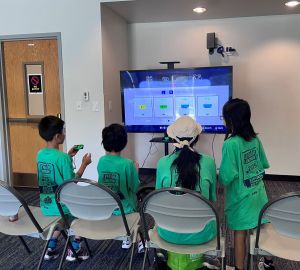 Four children facing away from the camera play video games on a large screen TV