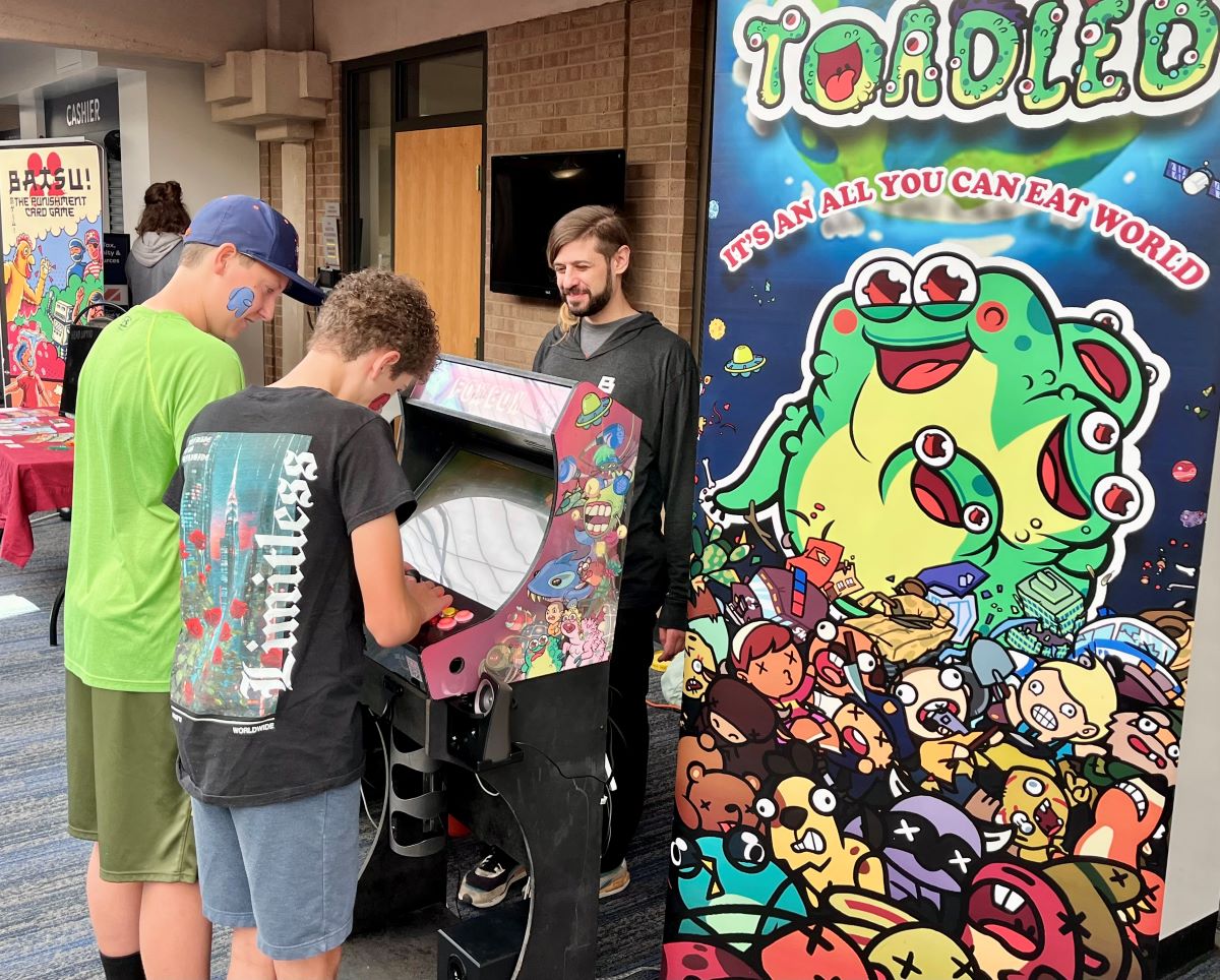 Two kids play at an arcade-style video game cabinet at LevelUp Longmont while an adult supervises