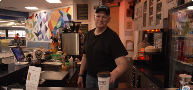 Christopher McCarthy, owner of Sinister Baker's Cafe, stands behind the counter offering a cup of coffee to the camera.