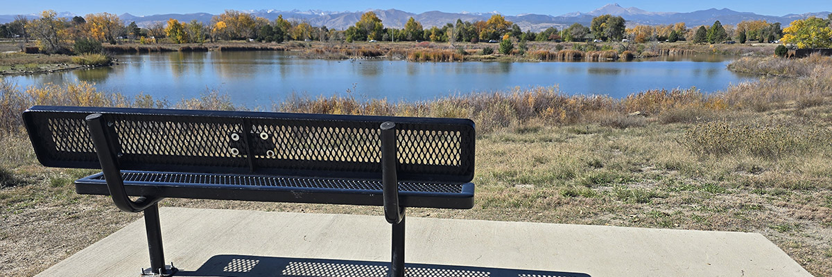 A black steel bend is mounted to a cement pad overlooking a lake with mountains and blue skies in the distance.