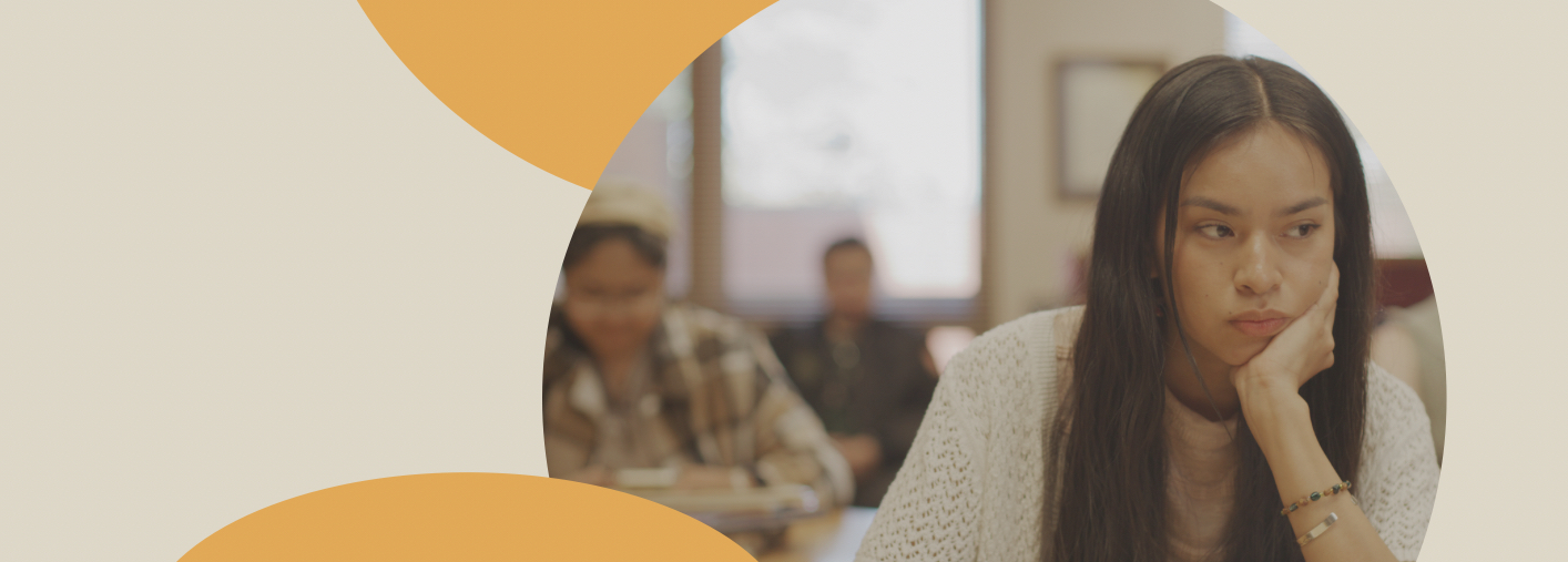 girl with long black hair in a classroom looking off to the left with a book on her desk