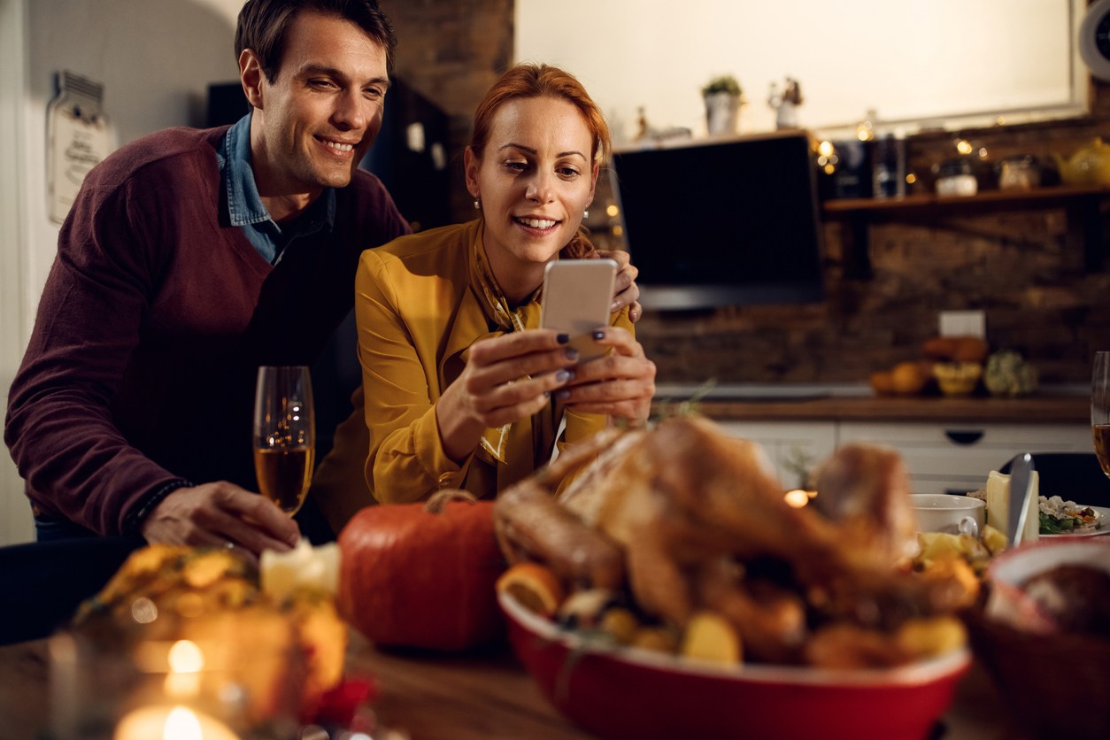 Happy couple on smartphone in kitchen with Thanksgiving feast